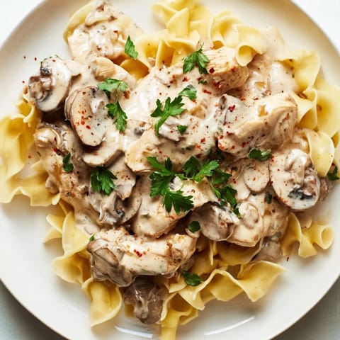 A close-up view of golden brown Chicken and Mushroom Stroganoff atop egg noodles, garnished with fresh parsley, ready for a satisfying weeknight dinner.