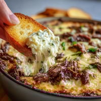 Golden, bubbly Philly Cheesesteak Dip baked in a dish with toasted crostini for dipping.