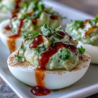 Creamy avocado deviled eggs topped with a spicy Sriracha drizzle, garnished with fresh cilantro for a vibrant appetizer.